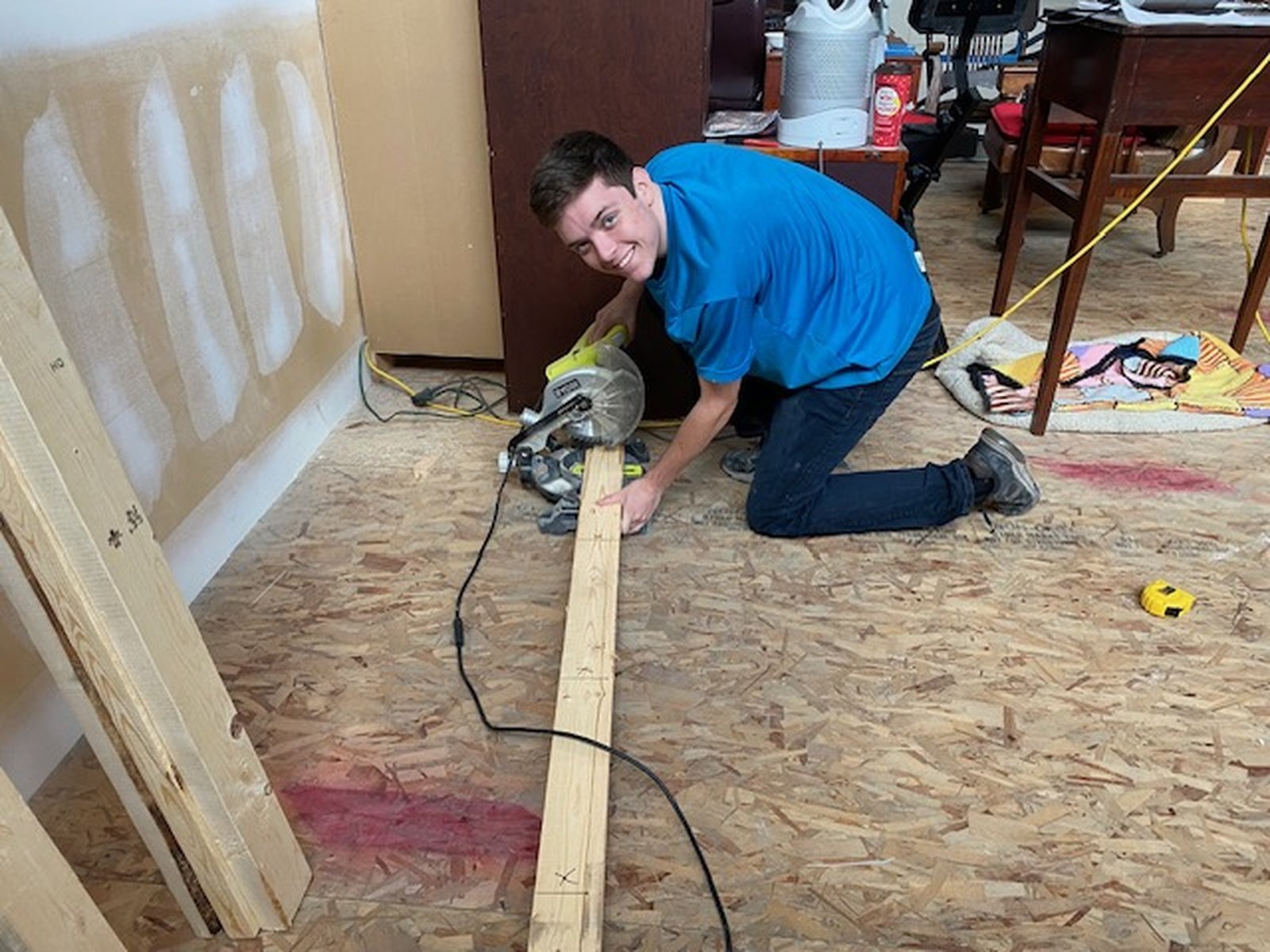 A Taylor Restoration Services technician kneeling on a bare subfloor, cutting a length of framing lumber with a mitre saw in a room stripped back for repair.