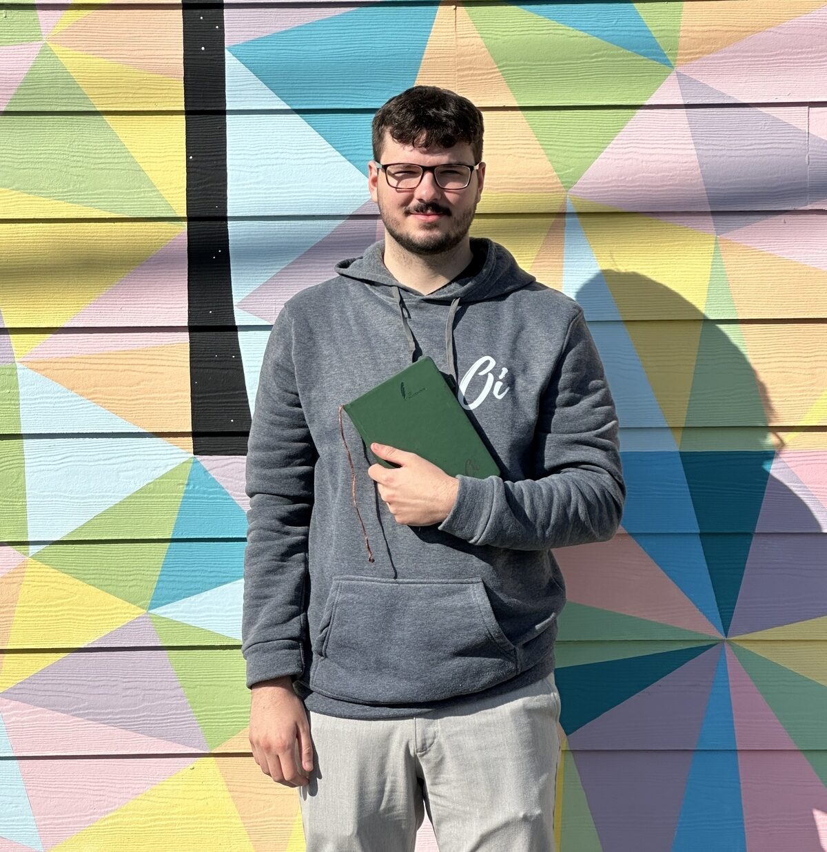 An Oi Marketing team member in an Oi-branded hoodie holding a green Oi-branded notebook, standing in front of a colorful geometric mural.