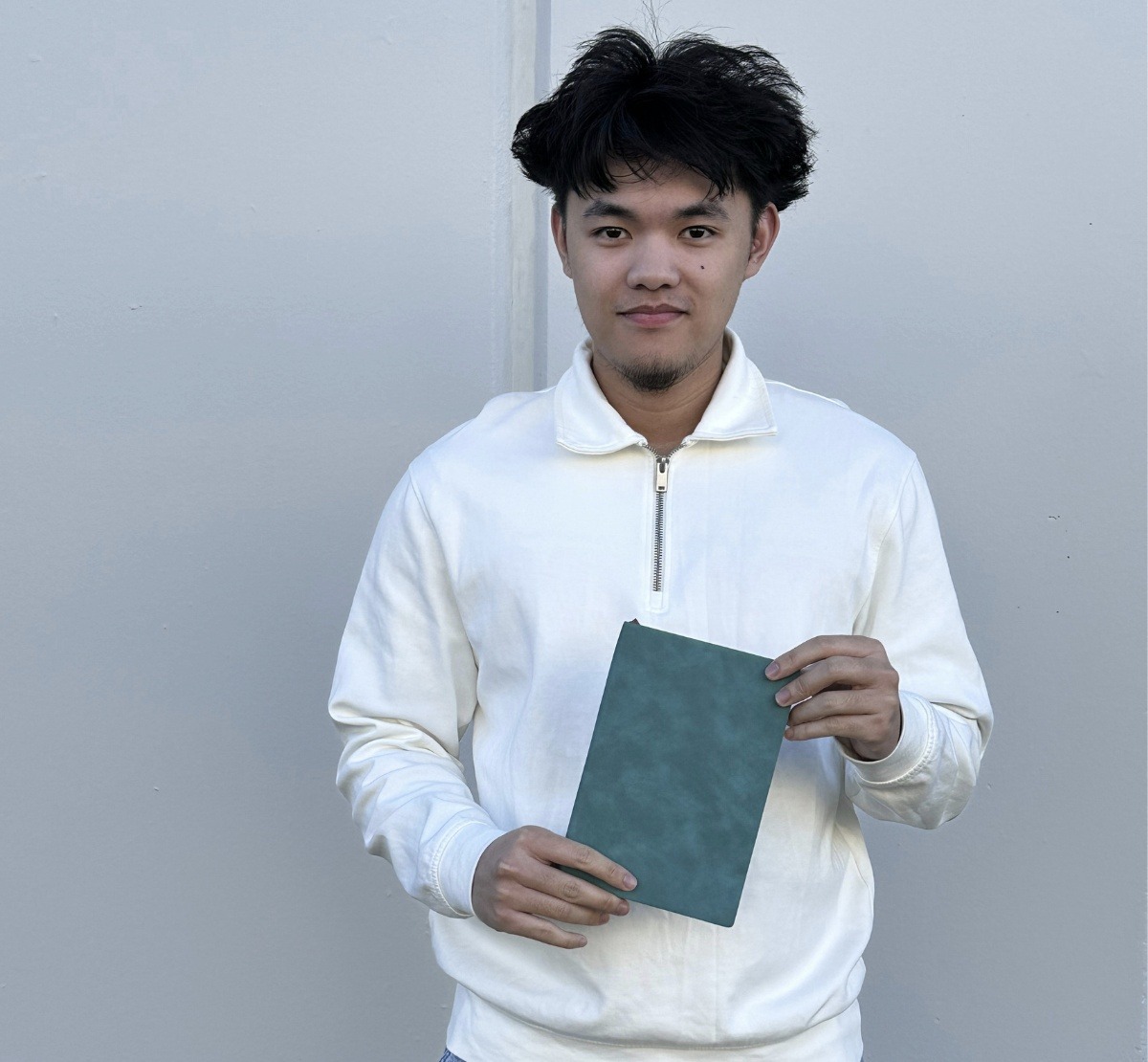 An Oi Marketing team member in a white pullover holding a teal Oi-branded notebook, standing against a plain wall.