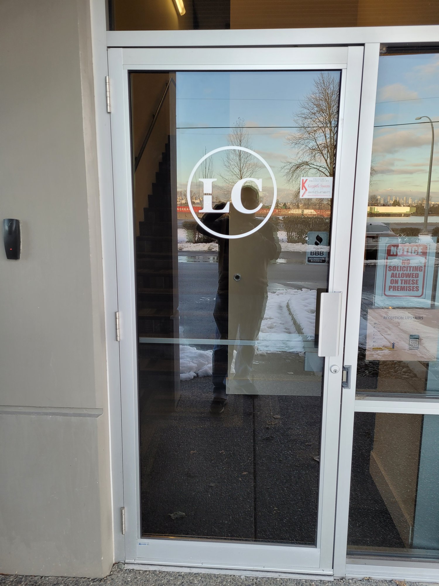 The glass entrance door at LabTest Certification's Delta office, marked with the white circular LC logo, with a BBB accredited business sticker and a no-soliciting notice on the adjacent window.