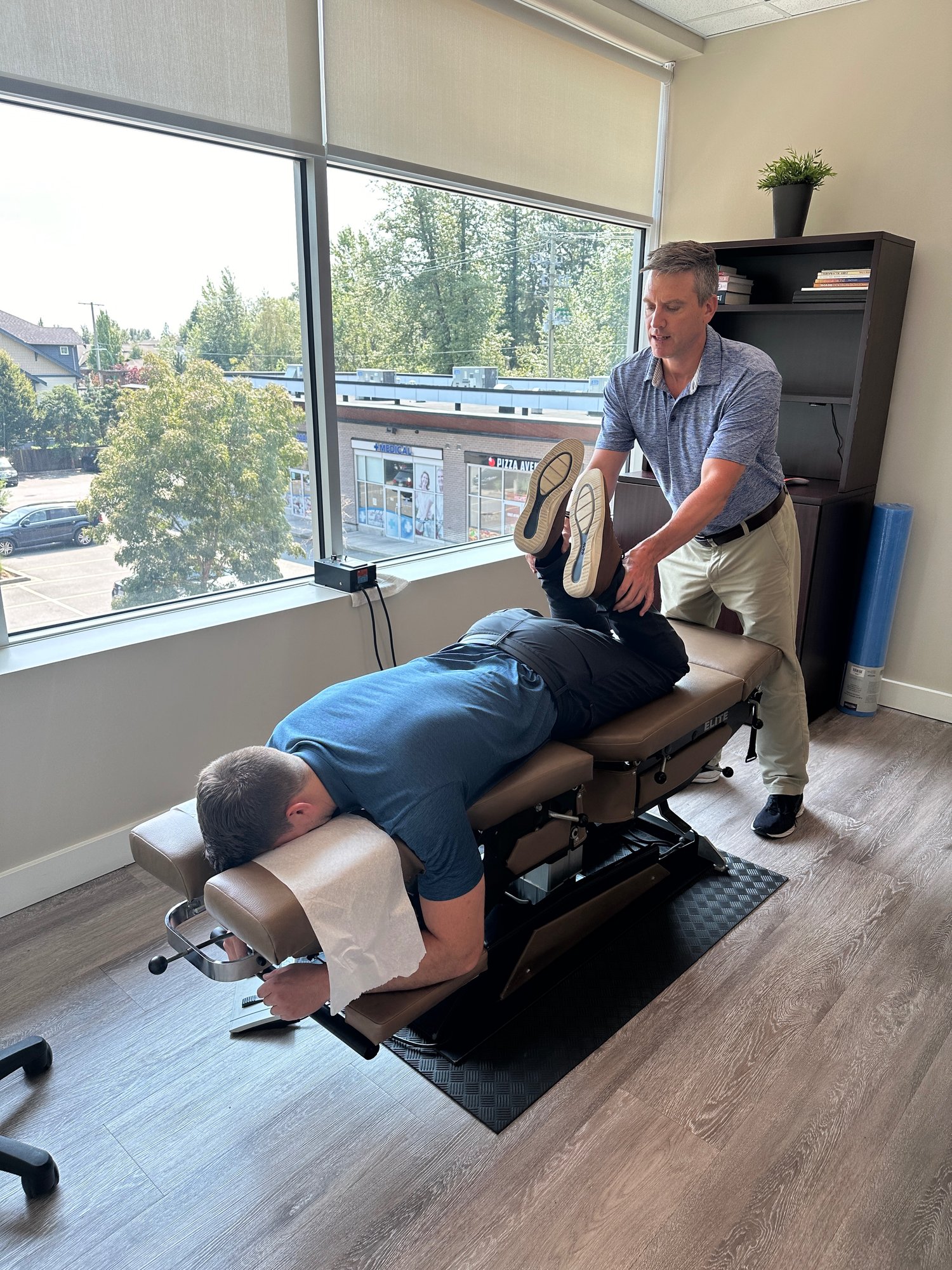 A chiropractor at Clayton Heights Chiropractic adjusting a patient who is lying face down on a drop table, beside a large window looking out over the street