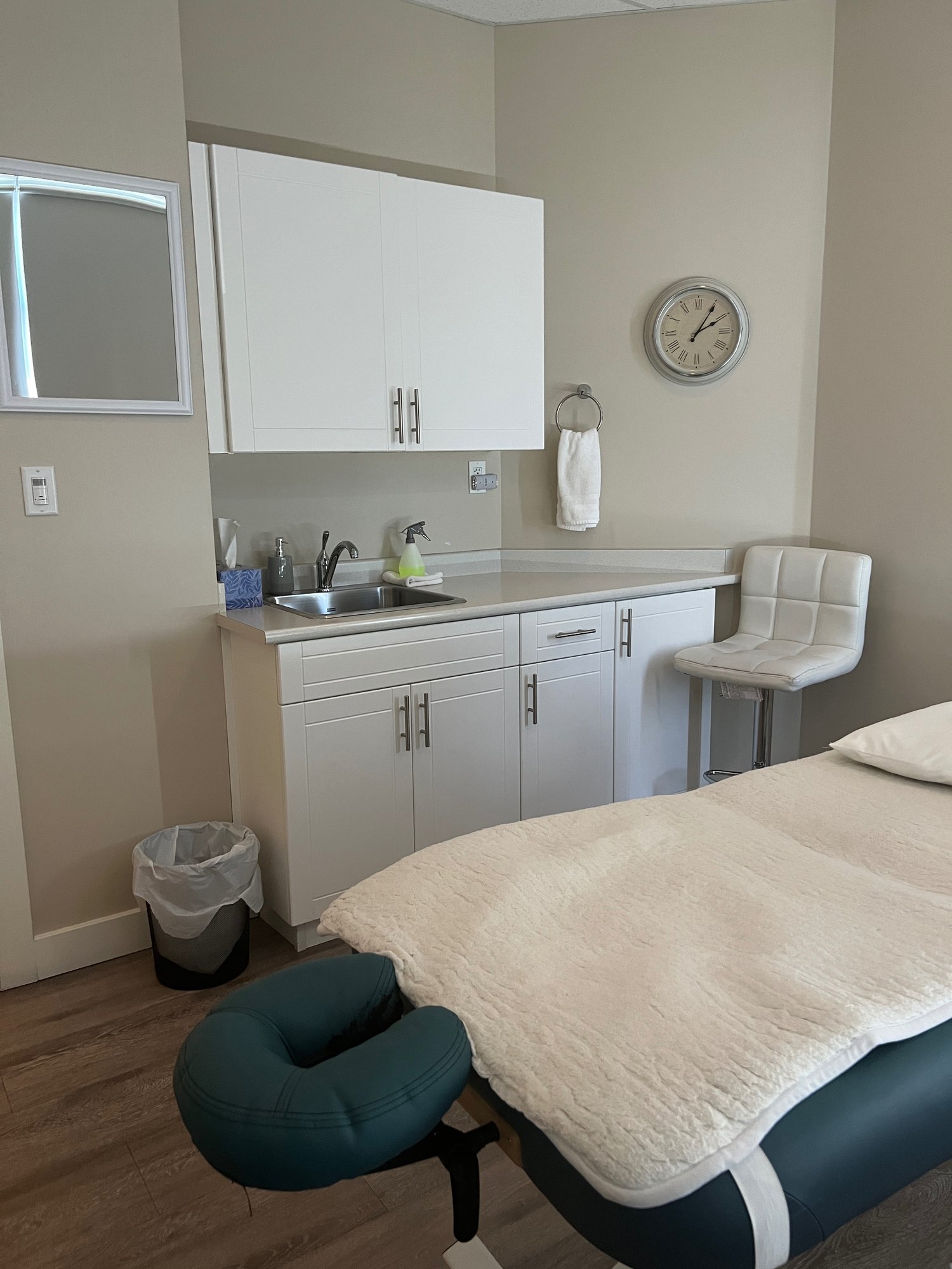 A treatment room at Clayton Heights Chiropractic with a teal massage table and face cradle, a wall clock, and a sink set into white cabinetry