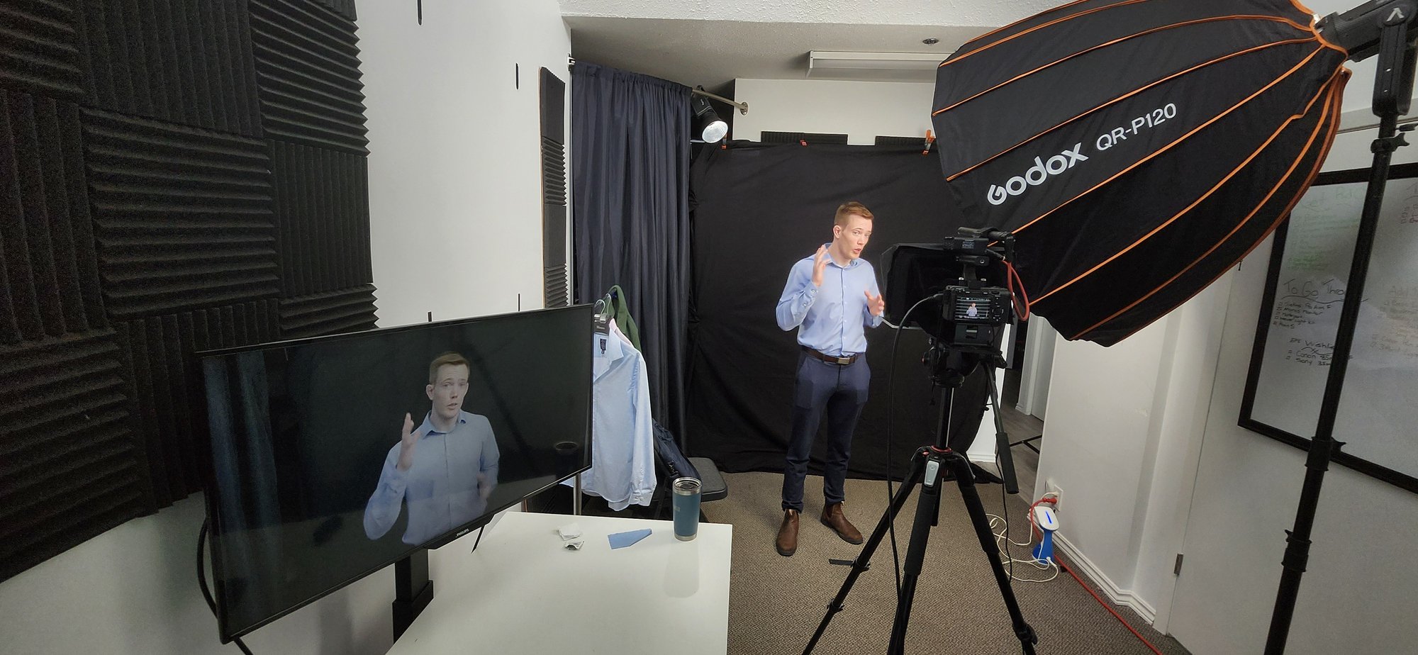 A presenter being filmed against a black backdrop in Dream It Creations' sound-treated studio, with a large softbox, a tripod-mounted camera and a monitor showing the live shot.