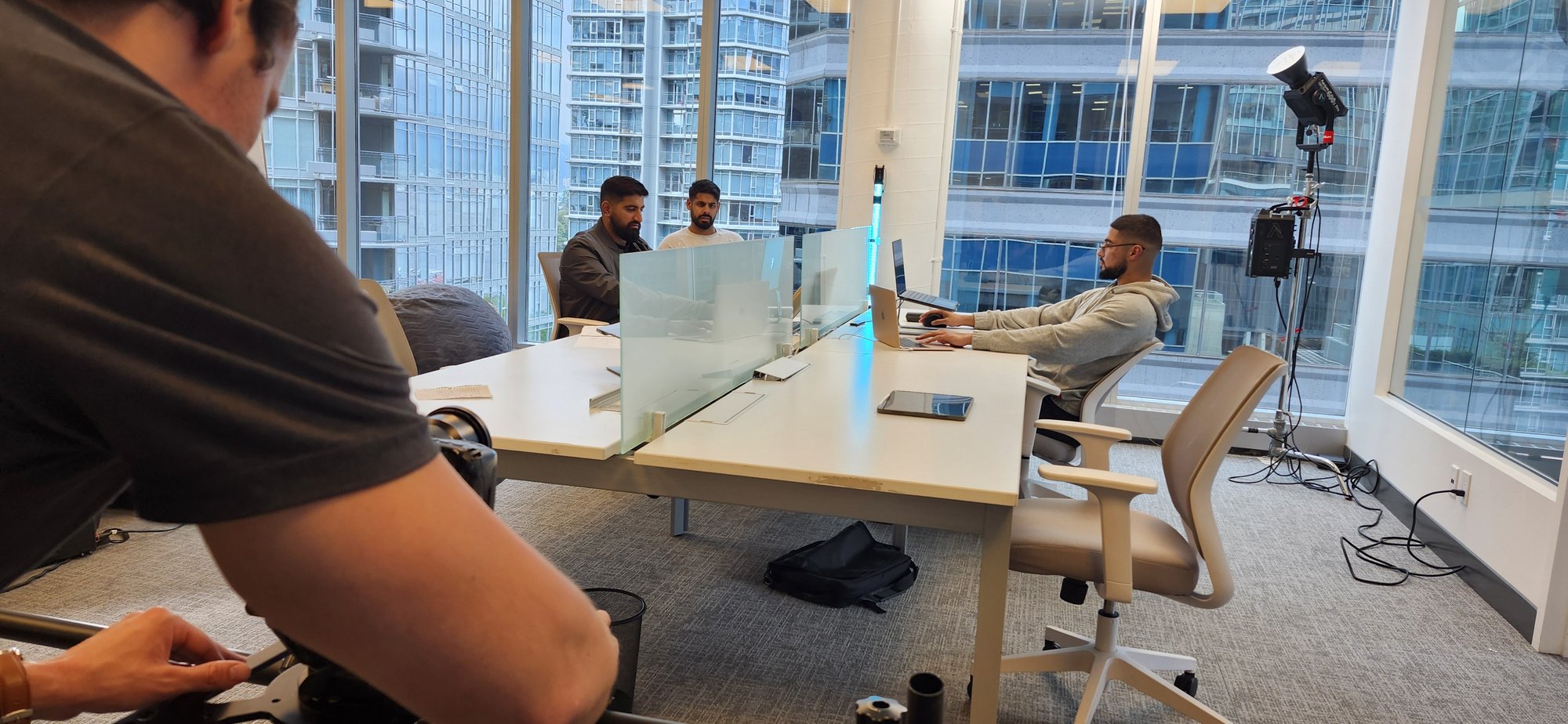 A Dream It Creations crew filming a corporate video in a glass-walled high-rise office, with a film light on a stand and staff working at white desks.