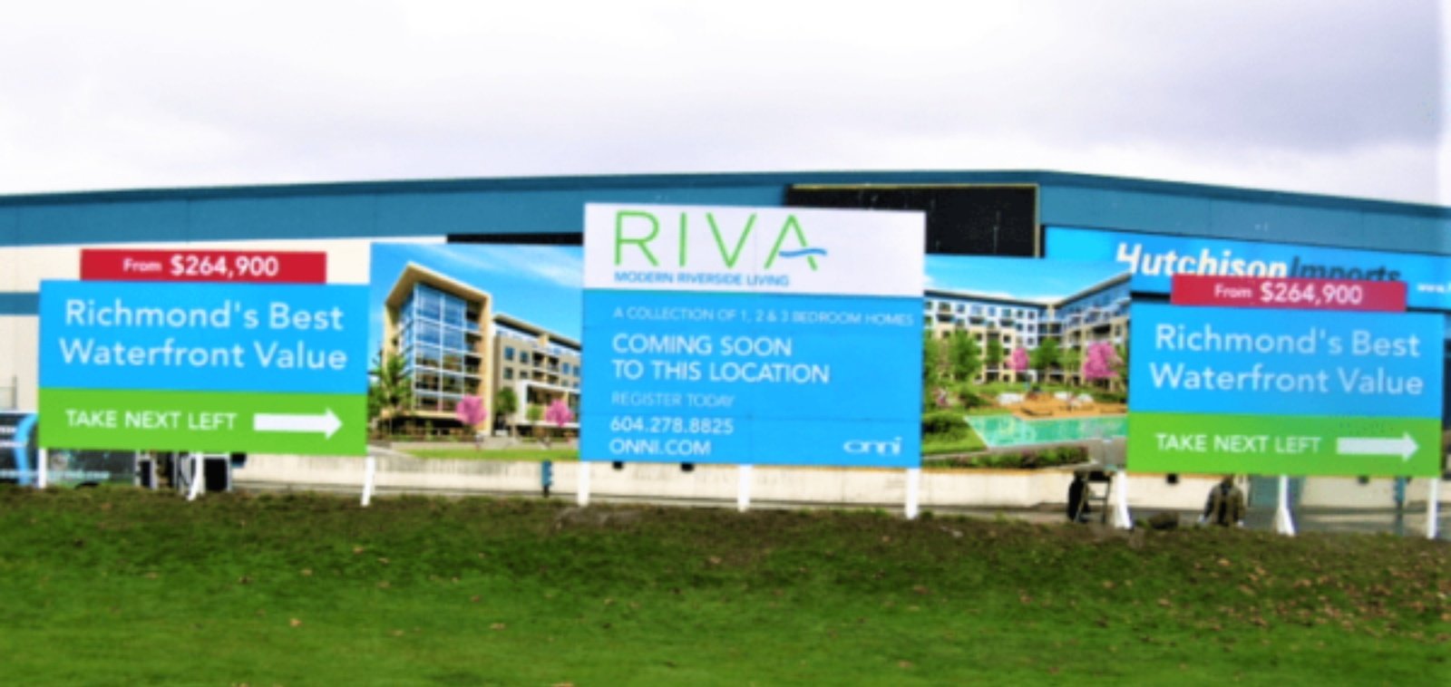 Large-format printed signage panels mounted along a grass verge in front of a building, reading "Richmond's Best Waterfront Value" and "From $264,900", flanking a RIVA by Onni "Coming Soon To This Location" development board.
