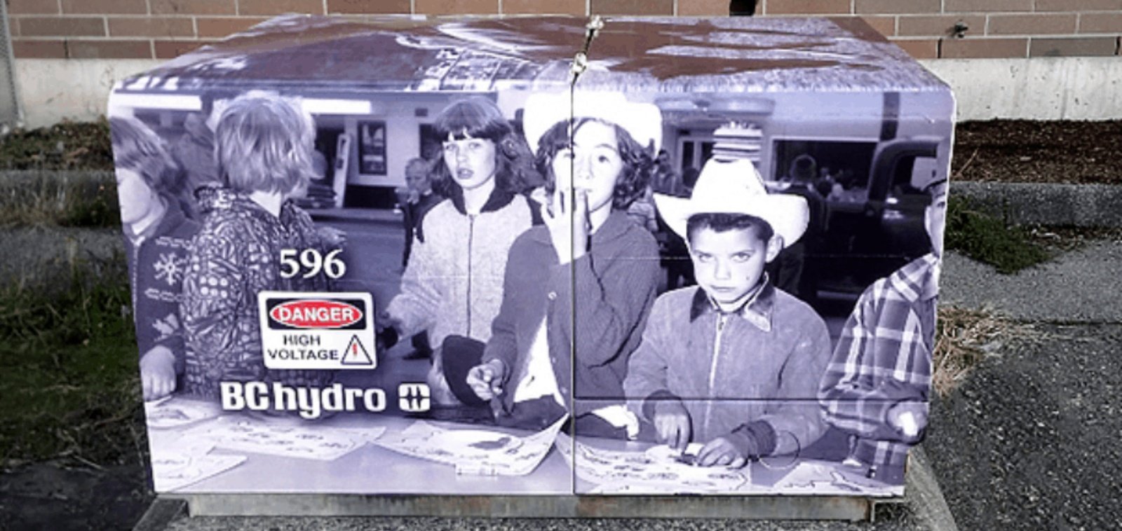 A BC Hydro electrical cabinet on a street corner wrapped in a printed black-and-white archival photograph of children gathered around a table, one boy in a white cowboy hat, with the utility's danger label and box number over the artwork.