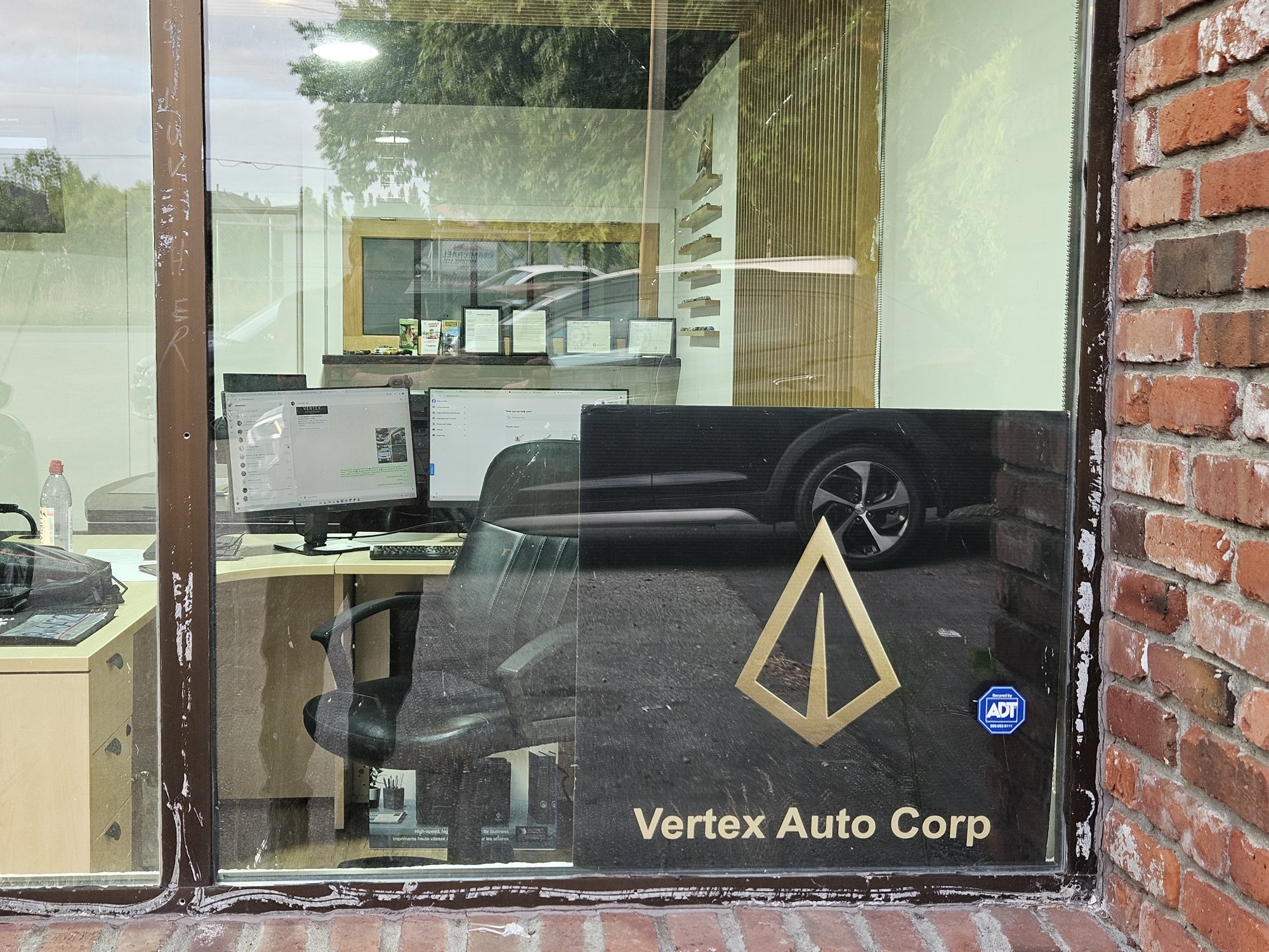 The Vertex Auto Corp storefront window in Langley, showing the company's gold arrow logo and name on a poster with the sales office desks visible through the glass