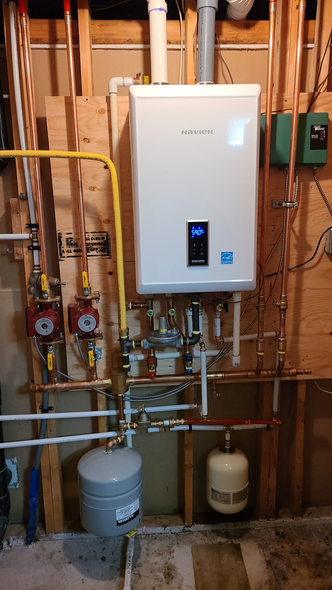 A newly installed Navien tankless combi boiler mounted on an open-stud basement wall, with copper supply lines, a yellow flexible gas line, two red circulator pumps and a pair of expansion tanks below.