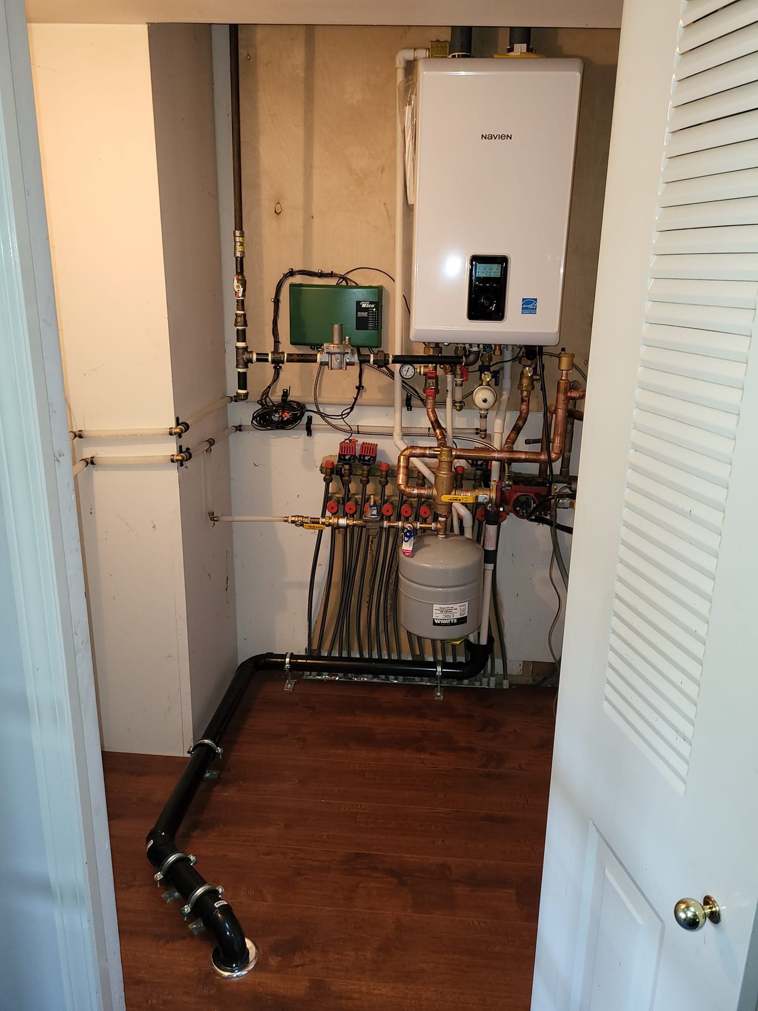 A completed Navien tankless water heater installation in a home utility closet, with copper piping, a radiant-heat zone manifold and an expansion tank above a black drain line running across the floor.