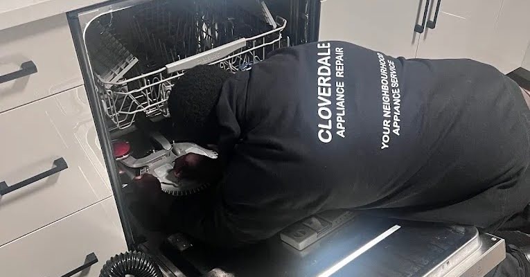 A Cloverdale Appliance Repair technician kneeling to repair the spray arm inside an open dishwasher.