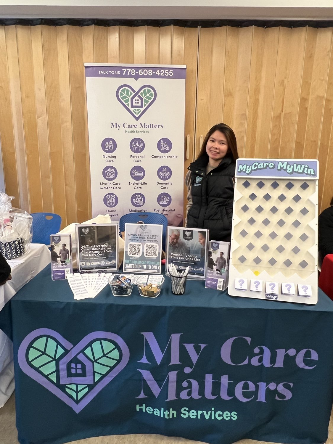 A My Care Matters Health Services team member at the company's information booth, with a teal branded table, a banner showing its phone number and care services, and a MyCare MyWin prize board alongside.