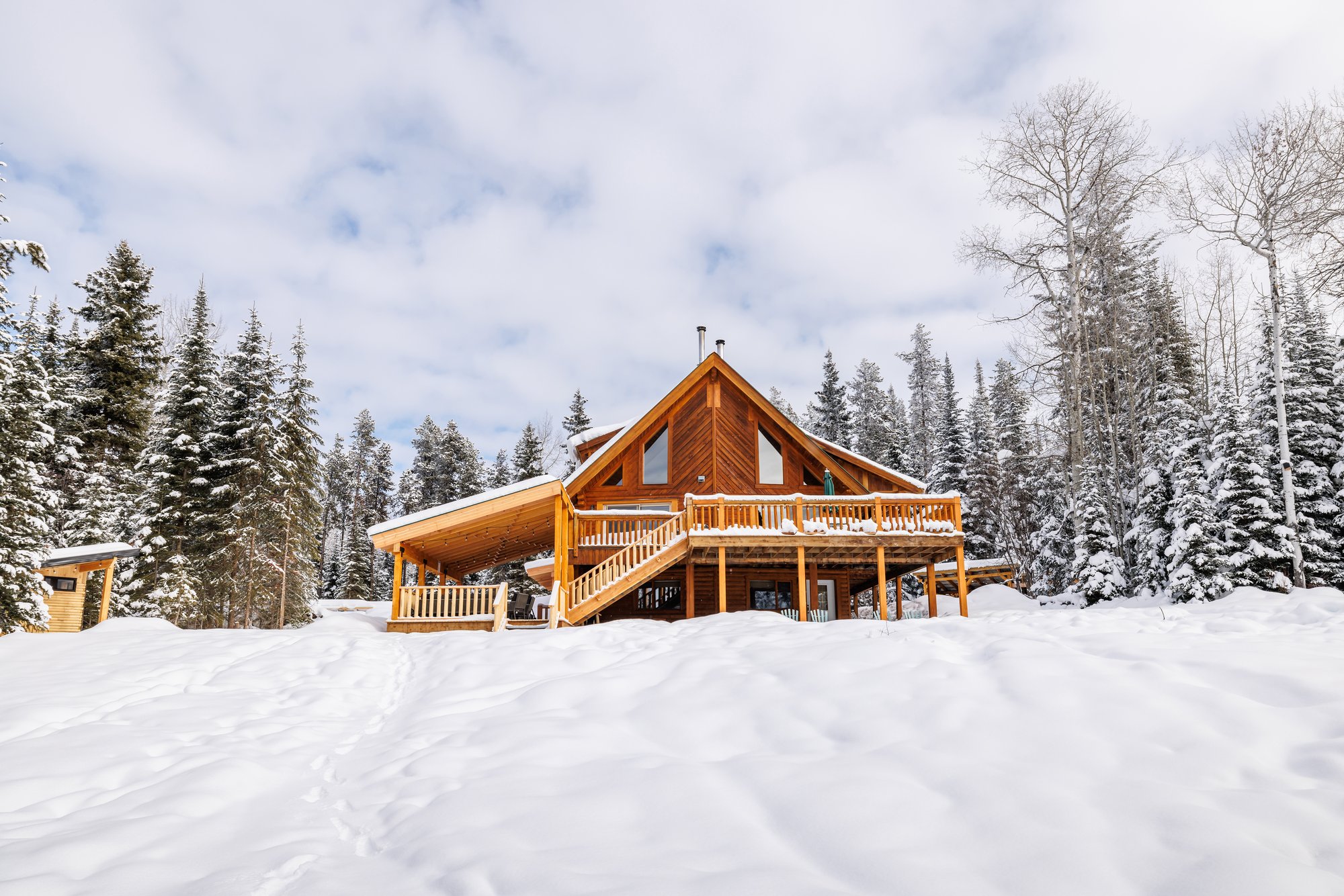 A cedar timber chalet with a railed deck, external staircase and covered lower patio standing in deep untouched snow, framed by snow-laden evergreens