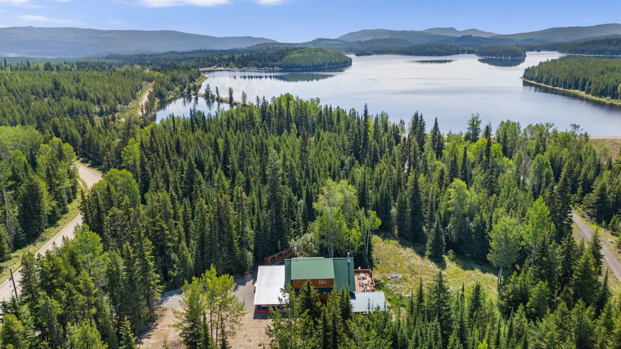 High aerial view of a green-roofed cabin in dense evergreen forest, with a long lake and forested hills stretching to the horizon behind it