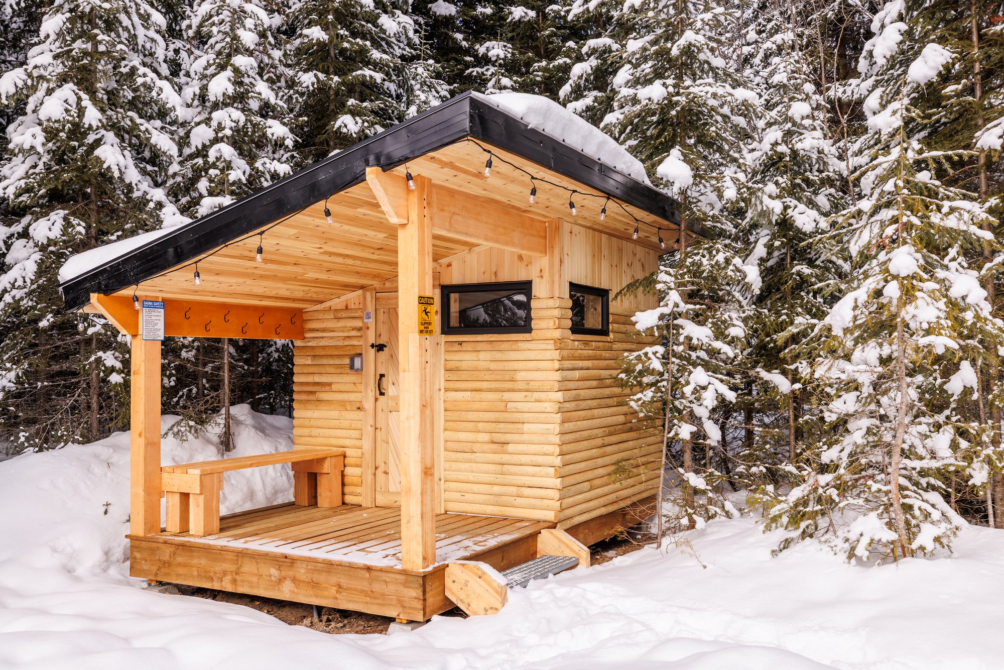 A small log-built outdoor sauna with a covered porch, bench, coat hooks and string lights, standing in snow at the edge of a snowy evergreen forest