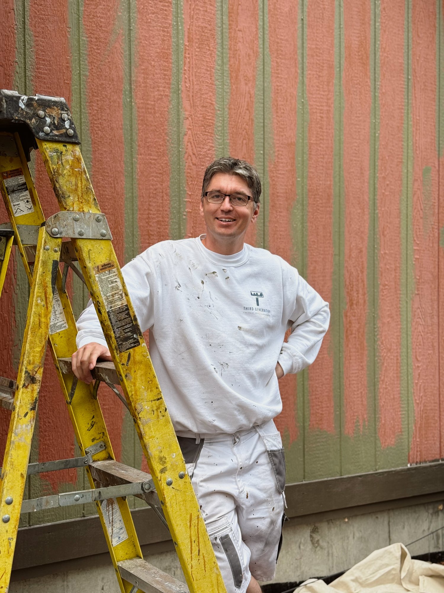 A smiling Third Generation Painters crew member in paint-spattered white work clothes, resting one hand on a yellow stepladder in front of wood siding that is part-way through being repainted from salmon to sage green.