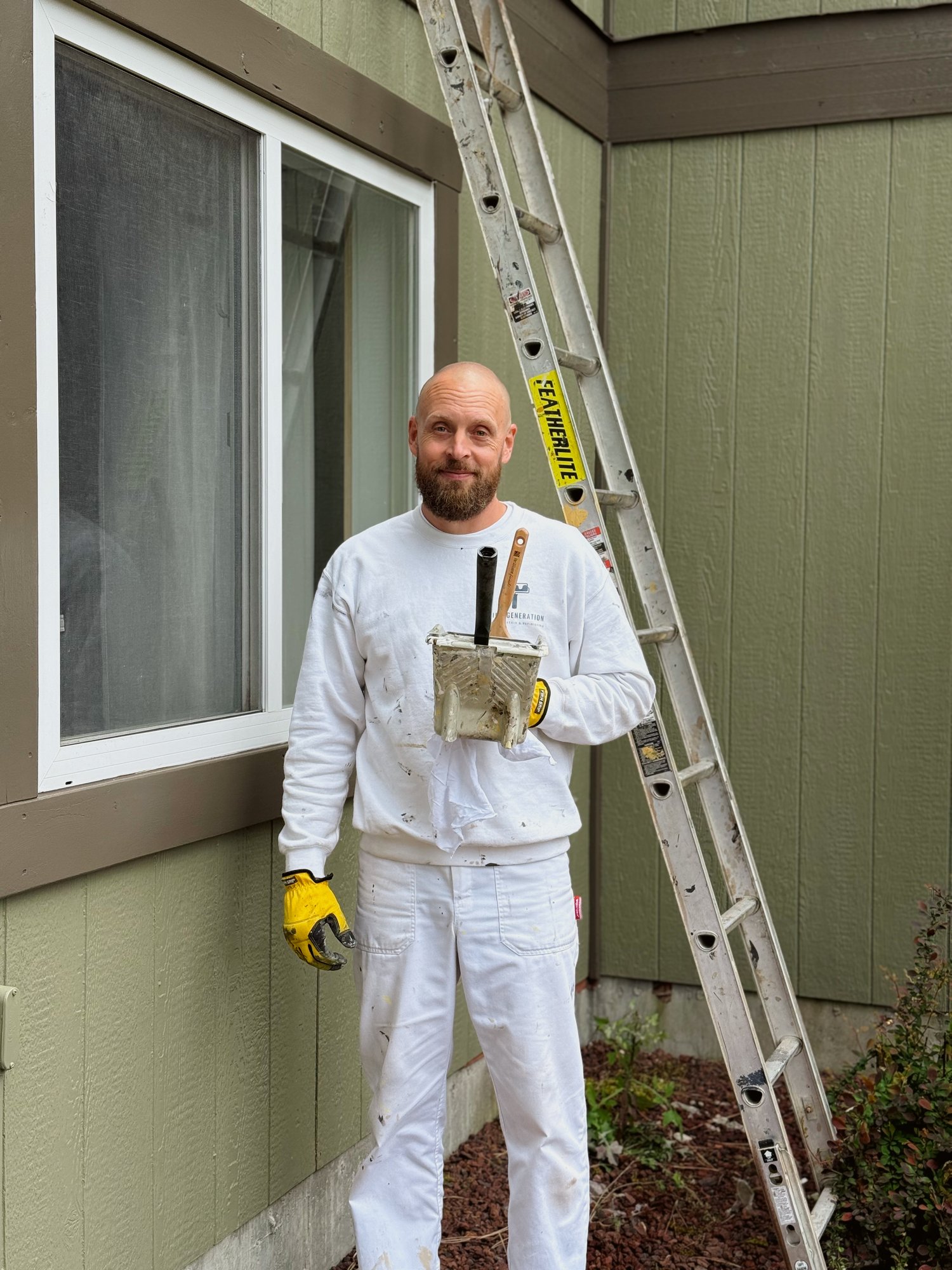 A bearded Third Generation Painters crew member in white work clothes holding a paint tray and brush, standing beside an extension ladder against a freshly painted sage-green house exterior.