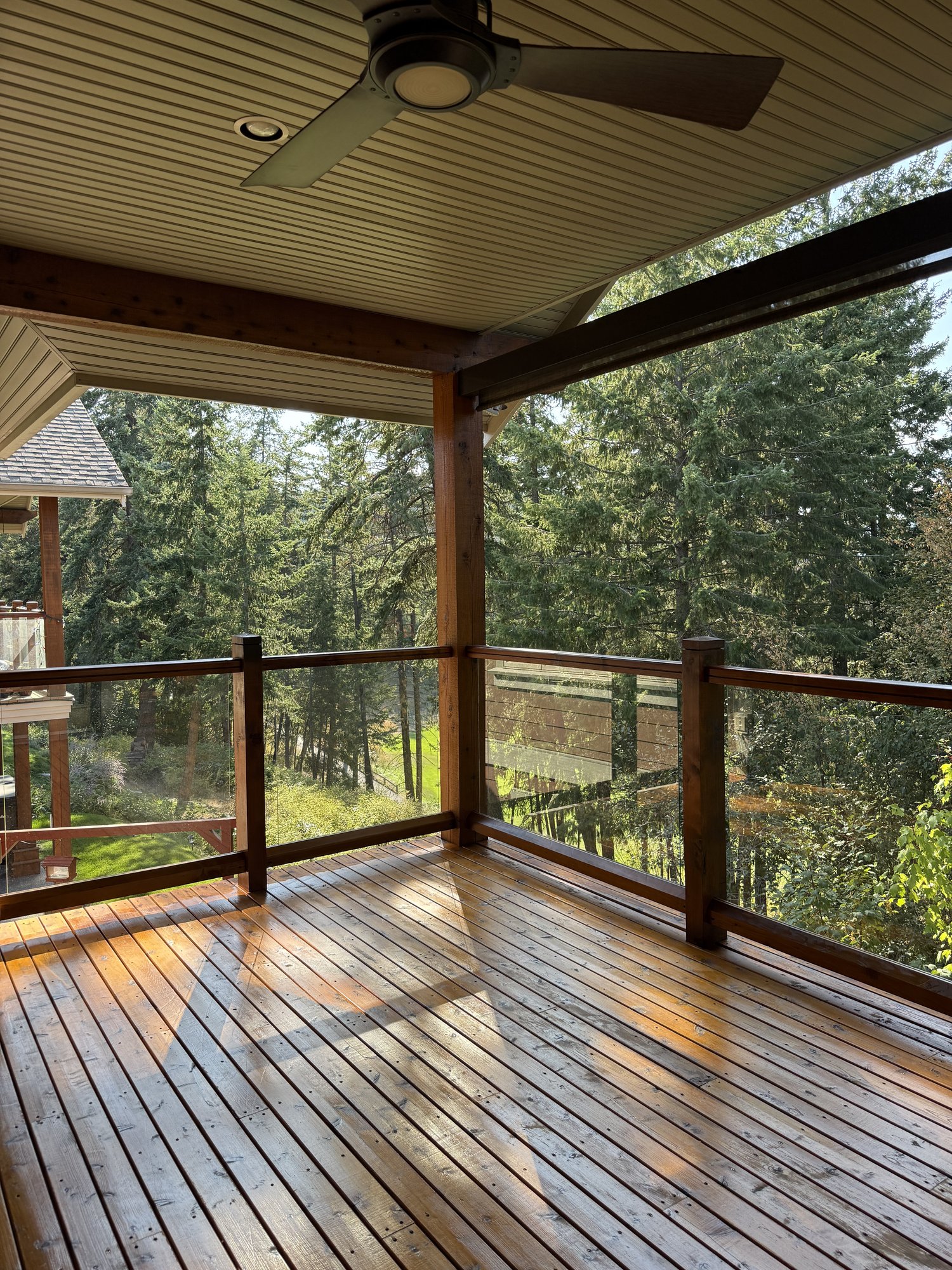 A covered wood deck with freshly stained decking, dark-stained posts and beams, and glass panel railings, looking out into evergreen forest.