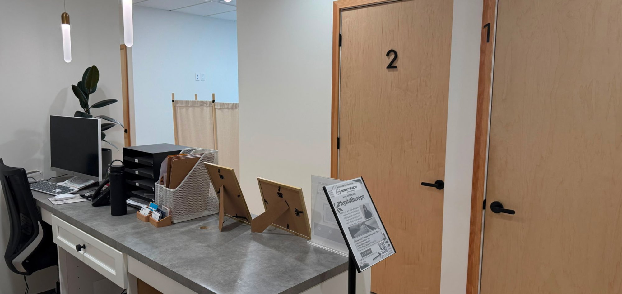 The front desk at Sonic Health Clinic, with a monitor, business card holders and a Sonic Health physiotherapy flyer on the grey counter, next to numbered treatment room doors.