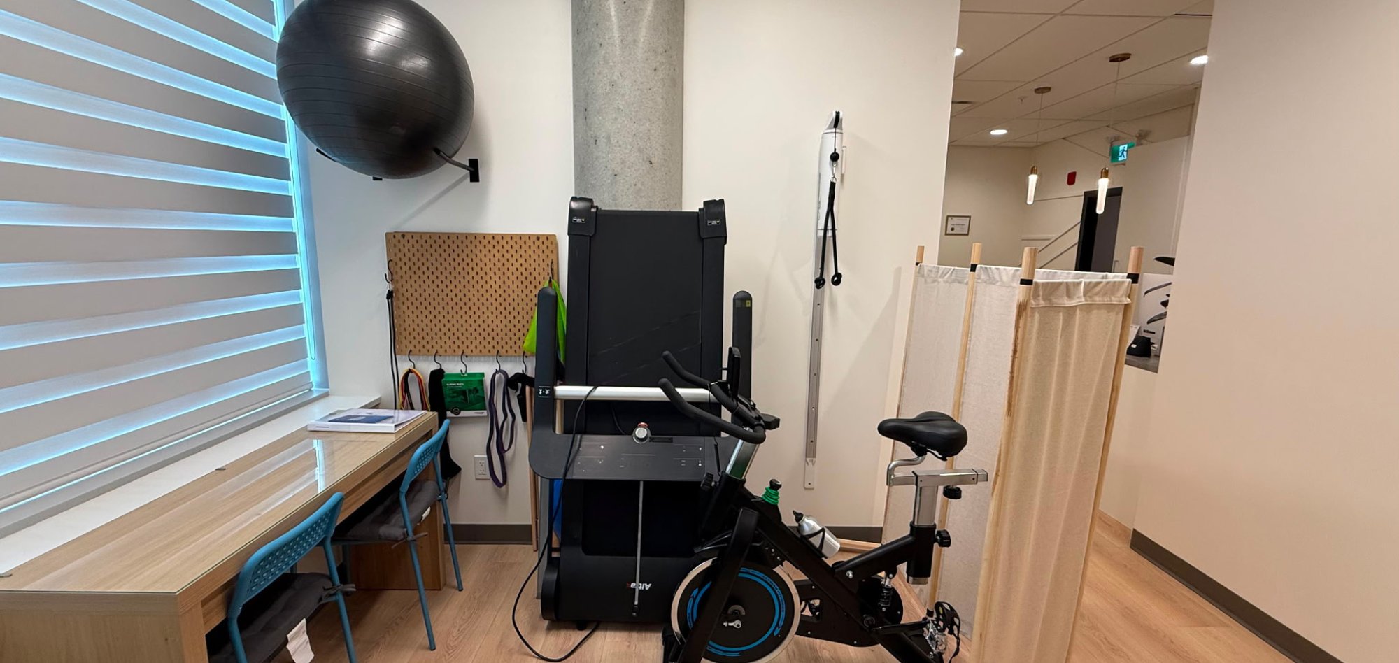 The rehabilitation and exercise area at Sonic Health Clinic, with a treadmill, a stationary spin bike, a large exercise ball mounted on the wall and resistance bands hanging from a pegboard beside a window.