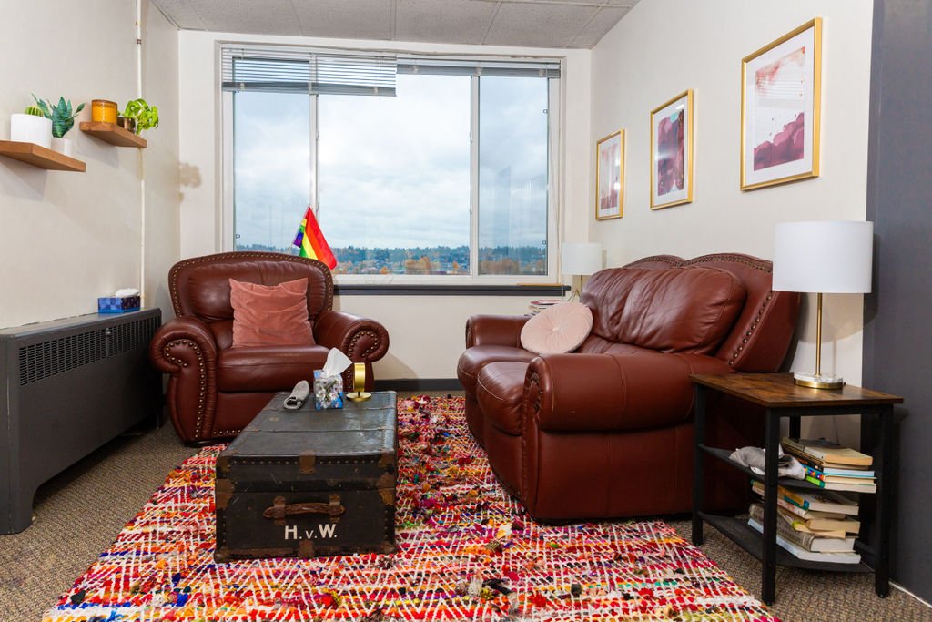 A counselling room with a brown leather armchair and loveseat facing each other across a vintage trunk, on a brightly coloured woven rug, with framed abstract art and a window overlooking treetops.