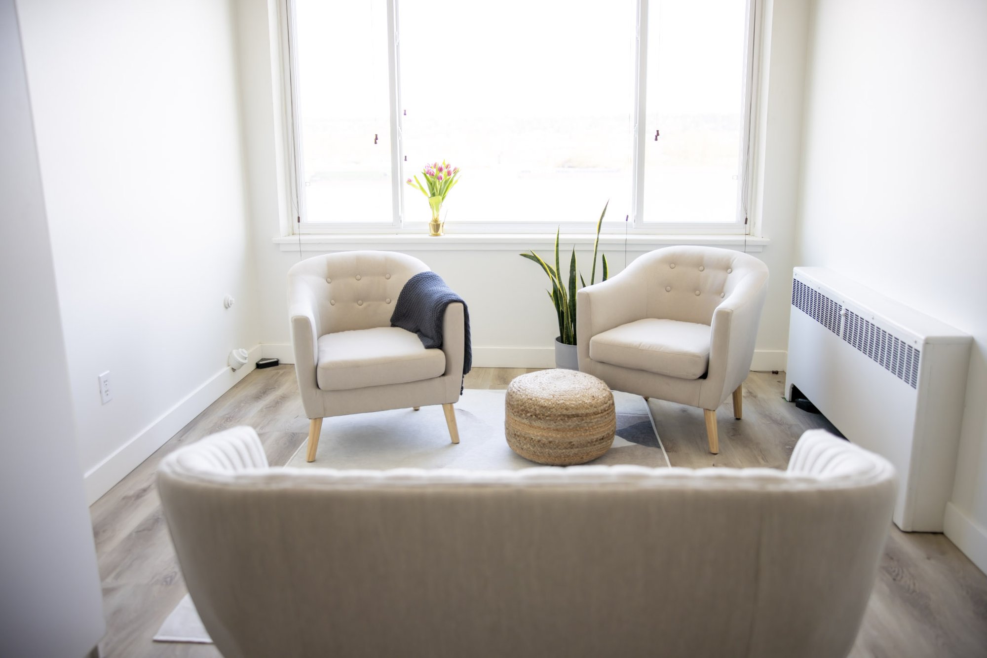 A bright counselling room with two cream armchairs and a woven pouf facing a sofa, beside tall windows with pink tulips on the sill.