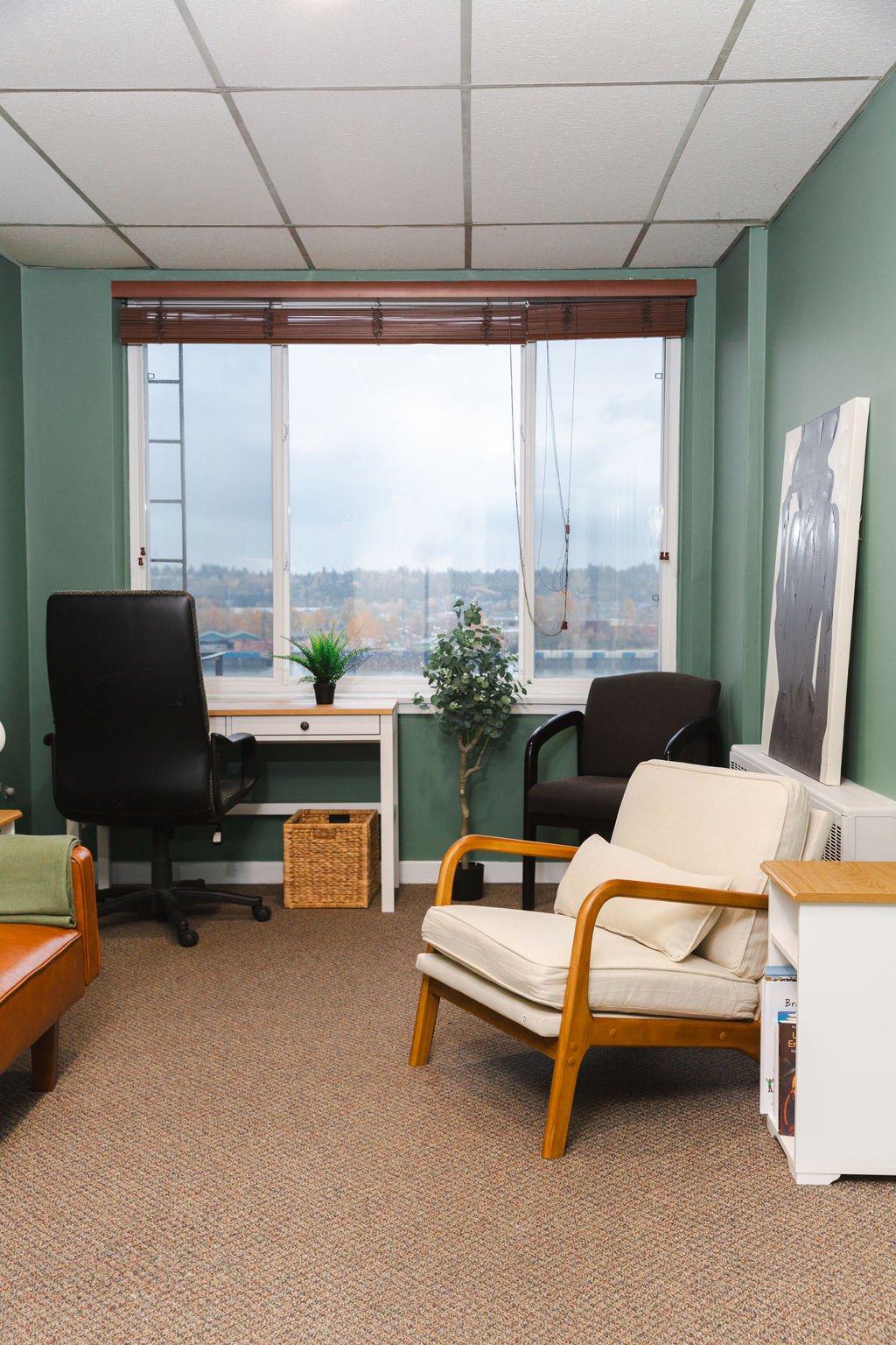 A sage-green counselling room with a wood-framed armchair and cream cushions, a white desk beneath a blind-shaded window, a leafy plant and a shelf of books.