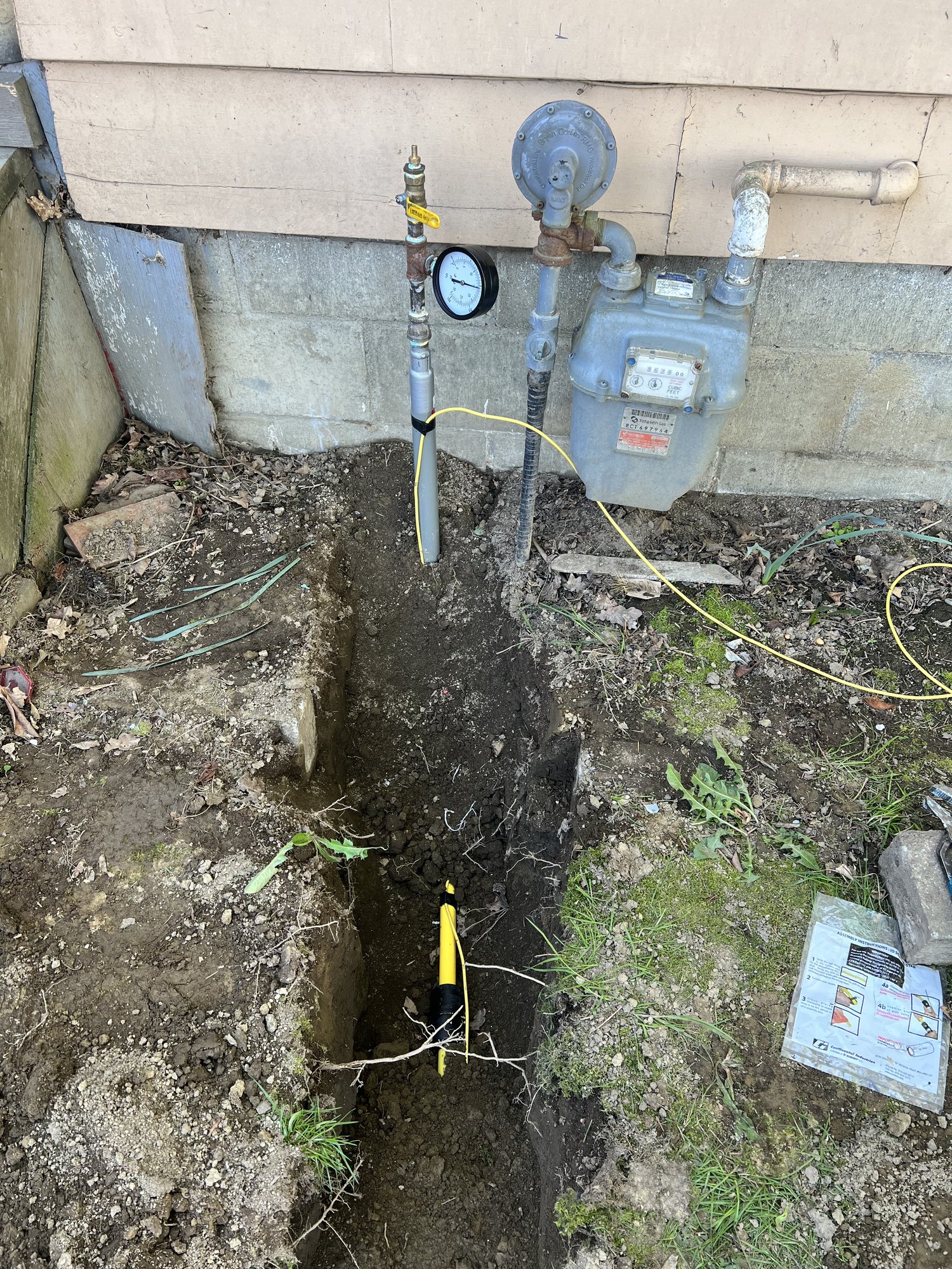 A new yellow polyethylene gas line laid in a hand-dug trench running up to a house gas meter, with a pressure test gauge fitted to the riser