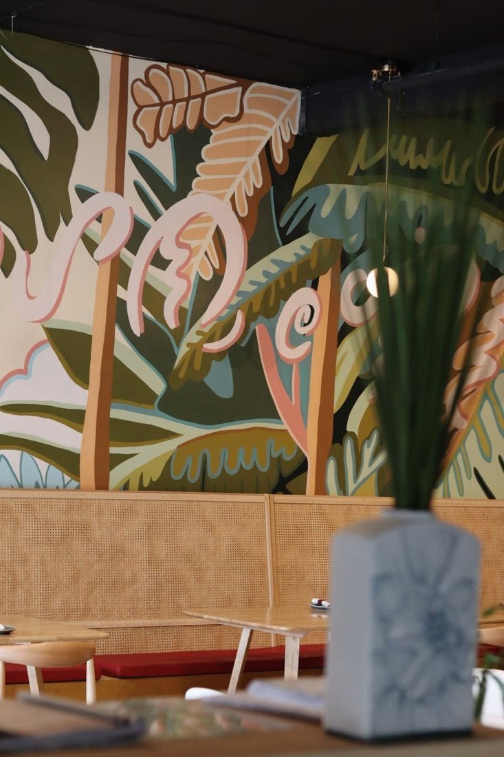 A hand-painted tropical leaf mural above a rattan banquette in the Halina dining room